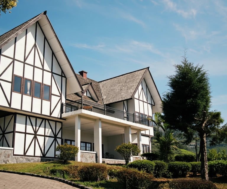 Rumah putih bergaya Eropa di bukit Puncak dengan atap cokelat, dikelilingi taman hijau.