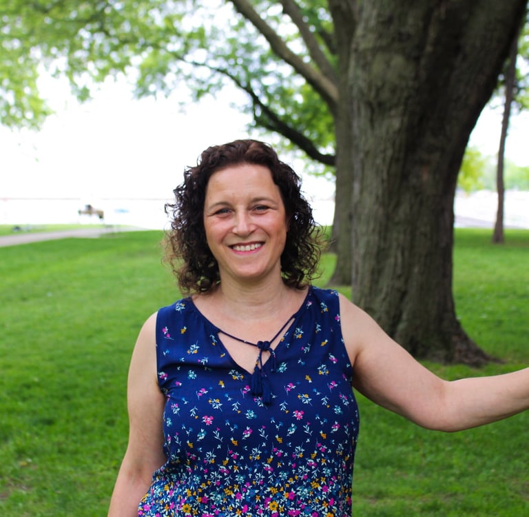 Picture of Natanya in a park, hand on a tree, water in the background.