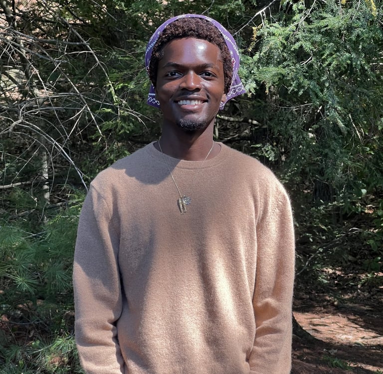 A smiling man wearing a beige sweater and purple bandana standing in a lush green forest.