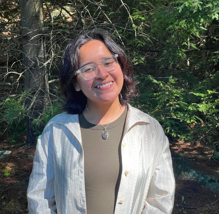 Smiling woman with glasses wearing a white shirt and olive top standing in a sunny evergreen forest.