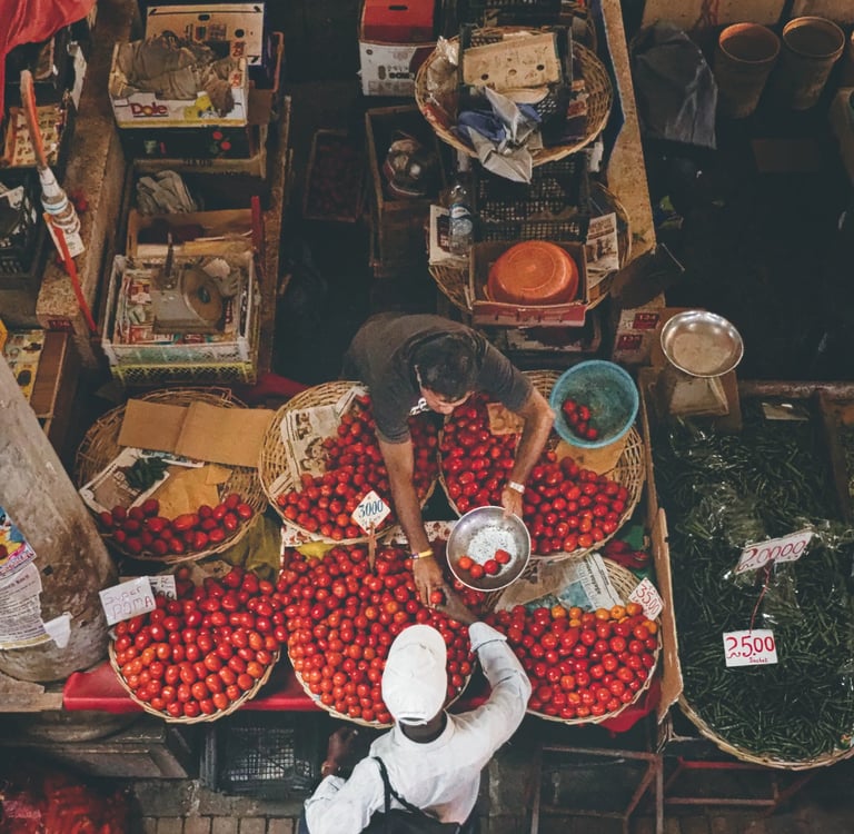 Overhead view of two men interacting at a market stall with baskets full of tomatoes, surrounded by various produce