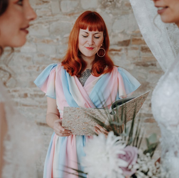 das queere Hochzeitspaar während der Trauung. die zwei Bräute sehen sich liebevoll an. die Rednerin blickt in ihre Rede-Mappe