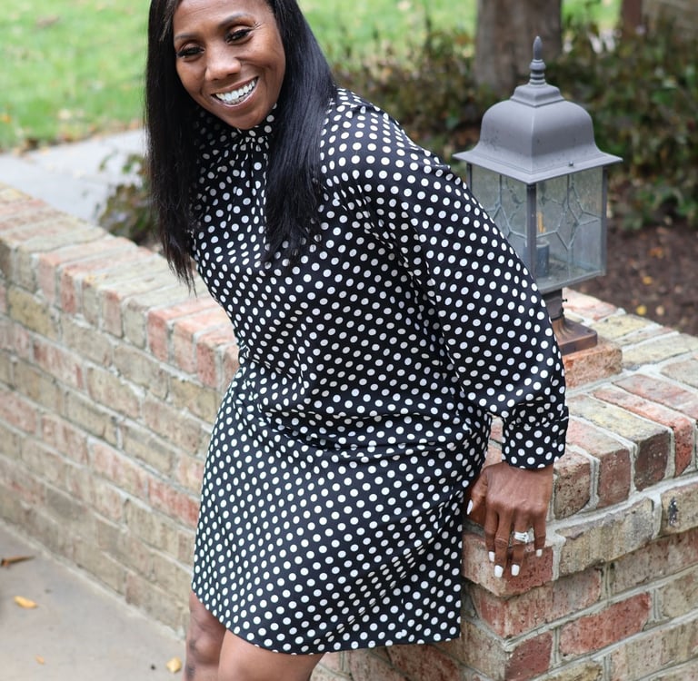 Smiling woman in a black and white polka dot dress posing against a brick wall outdoors.