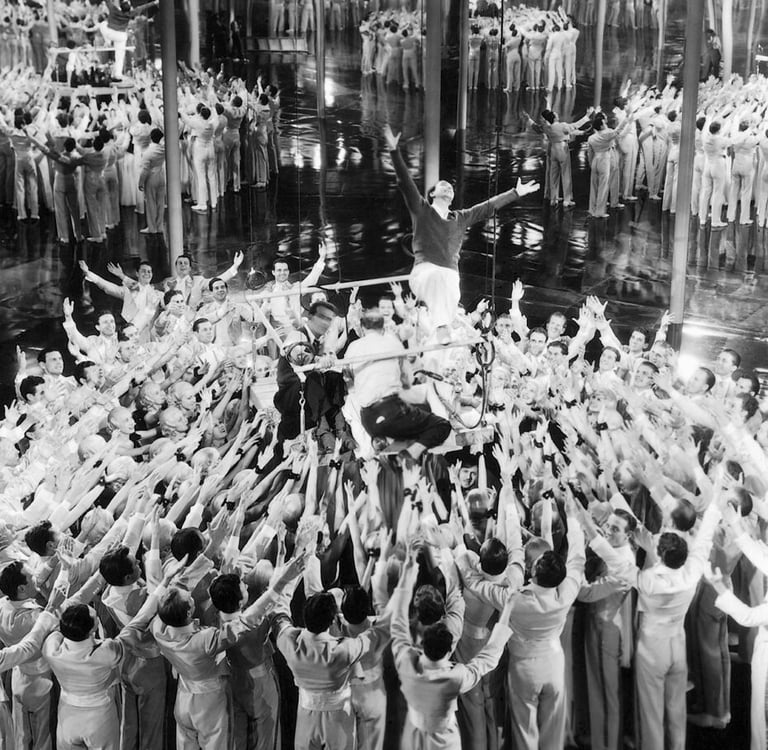 Busby Berkeley demonstrating arm movements to dancers on a mirrored stage platform.