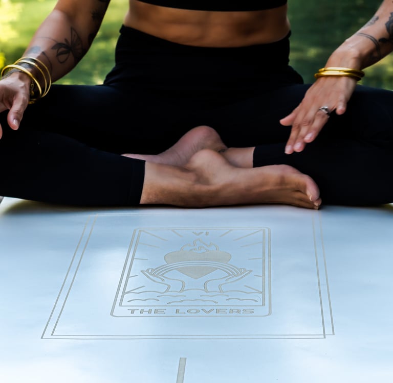 A woman sitting in a cross-legged yoga pose on a white yoga mat featuring The Lovers tarot card design.
