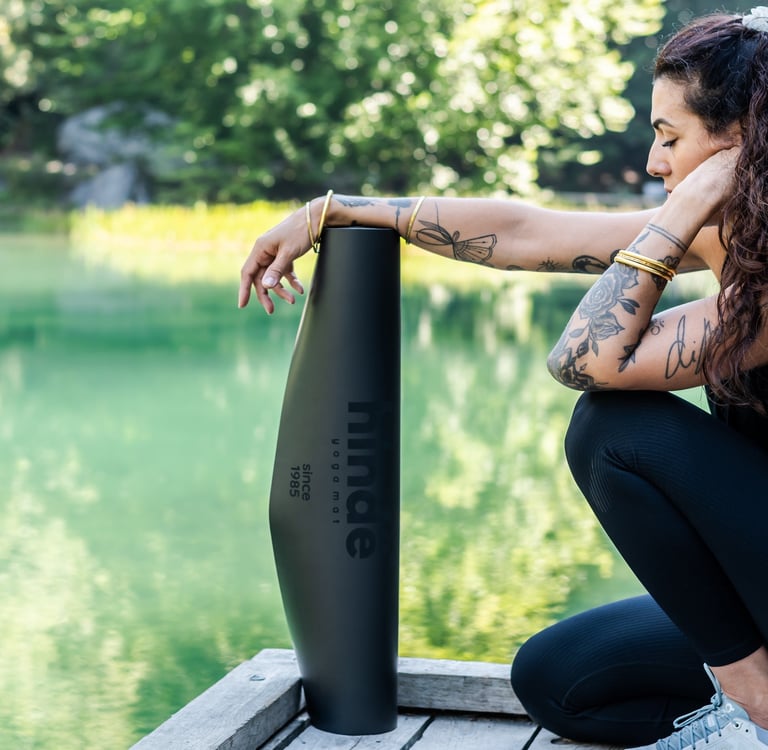 Tattooed woman posing with a black eco-friendly yoga mat on a wooden dock by a green lake.