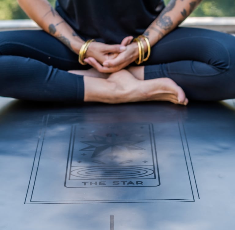 Tattooed woman sitting in a lotus pose on a black yoga mat featuring The Star tarot card design.