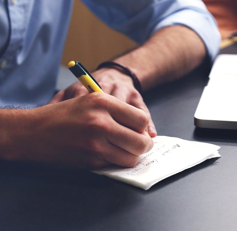 A person writing notes with a yellow pen on paper next to a laptop on a desk.