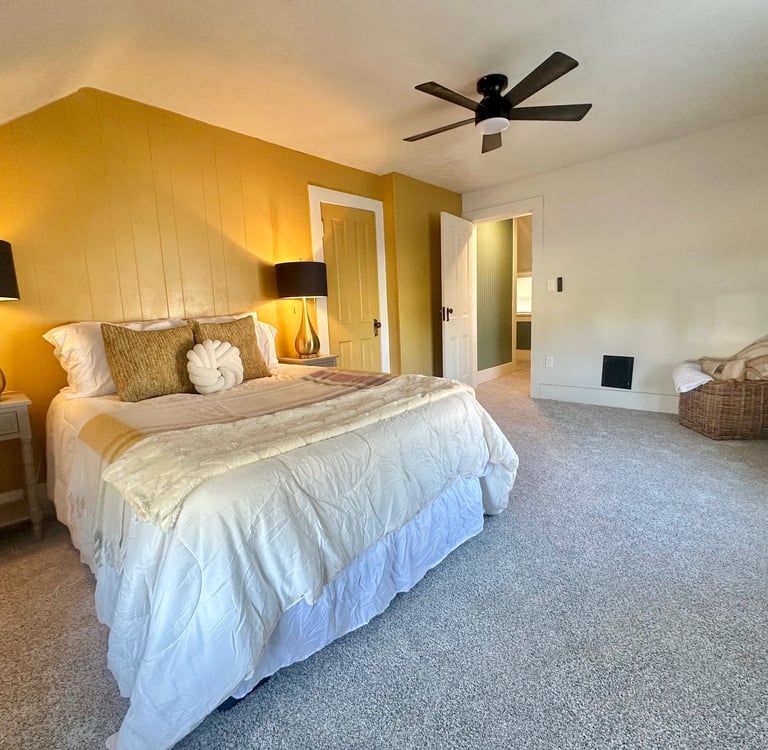 Cozy bedroom with a yellow accent wall, white bedding, gold lamps, and a black ceiling fan.