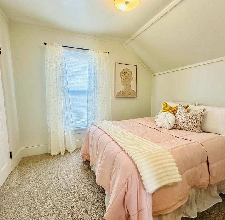 Cozy bedroom with pink bedding, decorative pillows, and white curtains near a window with natural light.