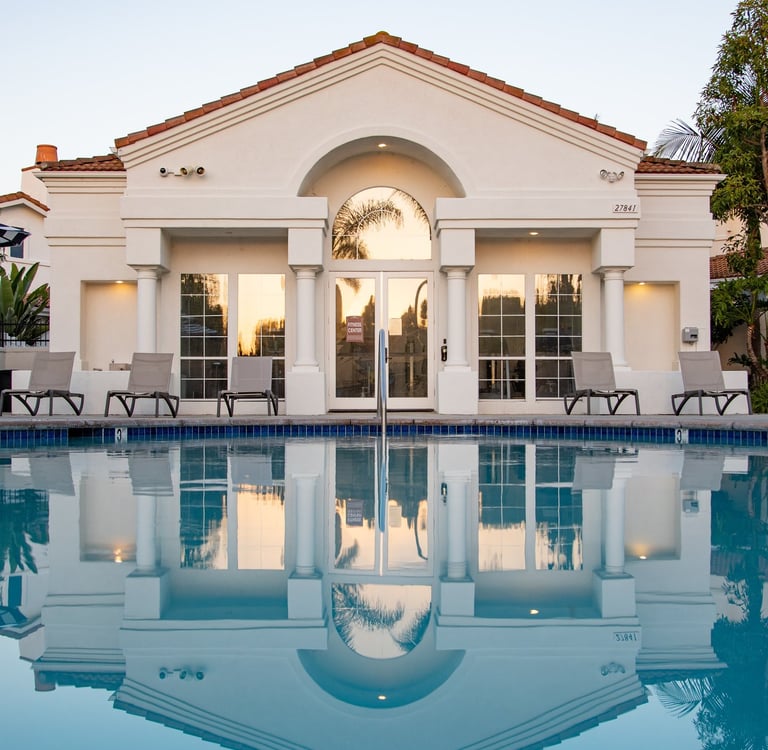Off-white facade of luxury building with pillar architecture. A pool in front and deckchairs.