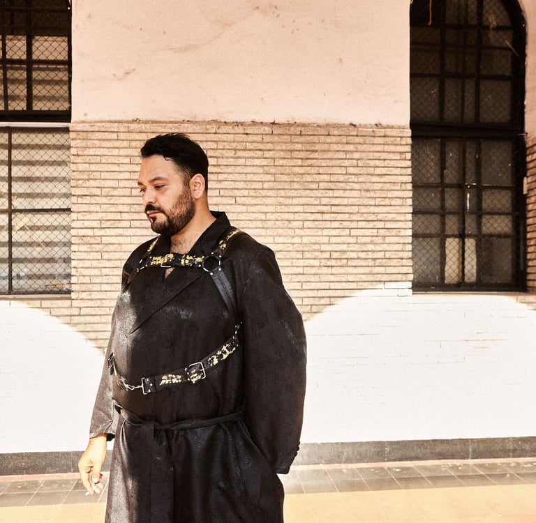 A man in a black avant-garde robe and leather body harness stands before a brick wall with windows.