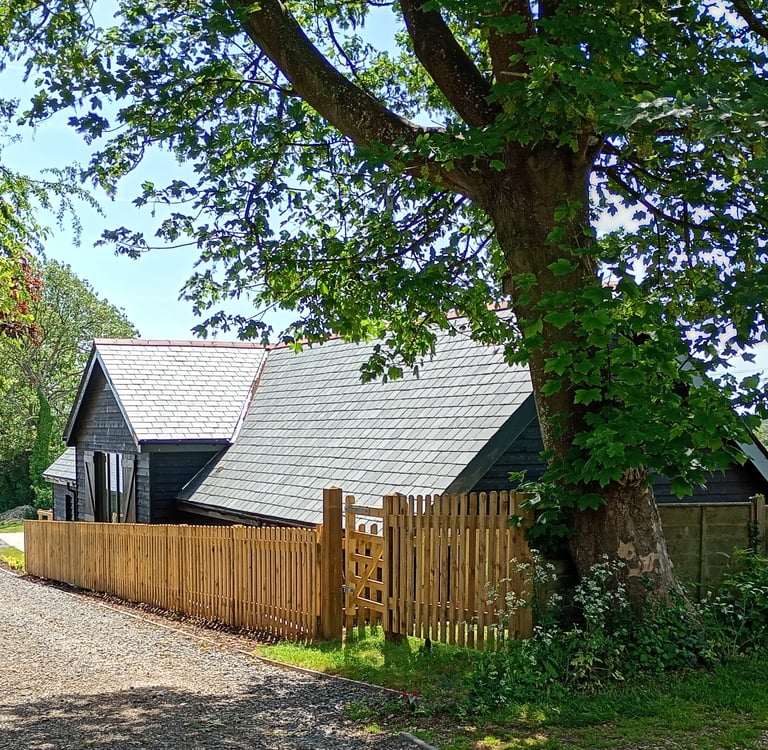 A black timber barn conversion with a slate roof behind a wooden picket fence and large tree.