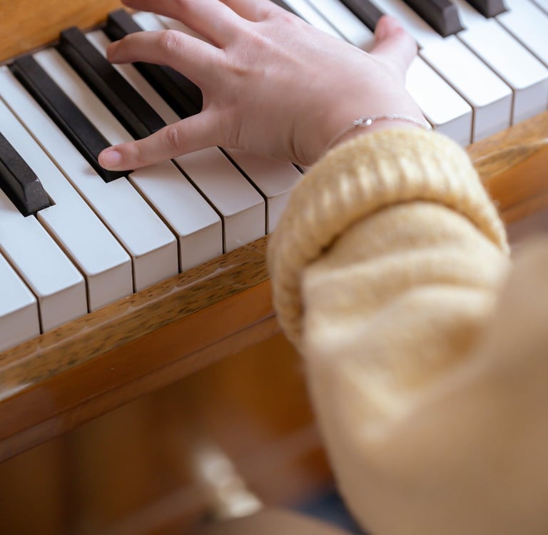 A person wearing a yellow sweater plays a wooden upright piano during a music lesson.