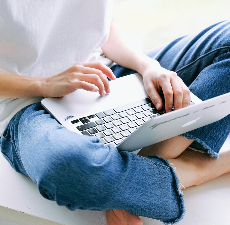 A person in blue jeans sitting cross-legged while typing on a laptop computer.