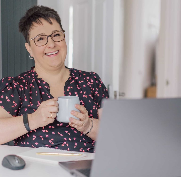 Smiling woman with short hair holding a coffee mug while working on her laptop at home.