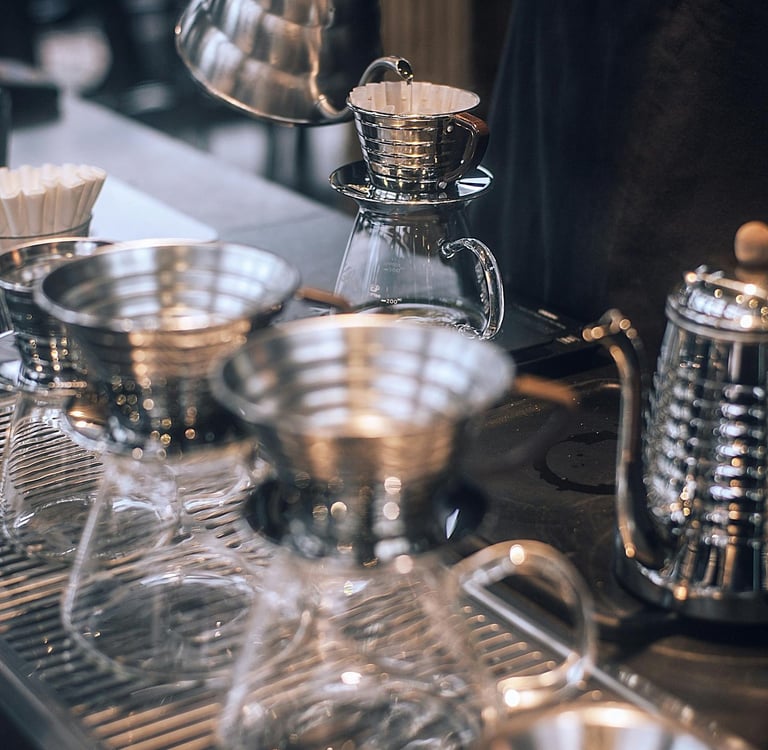 Barista pouring hot water from a gooseneck kettle into a stainless steel pour-over coffee dripper.