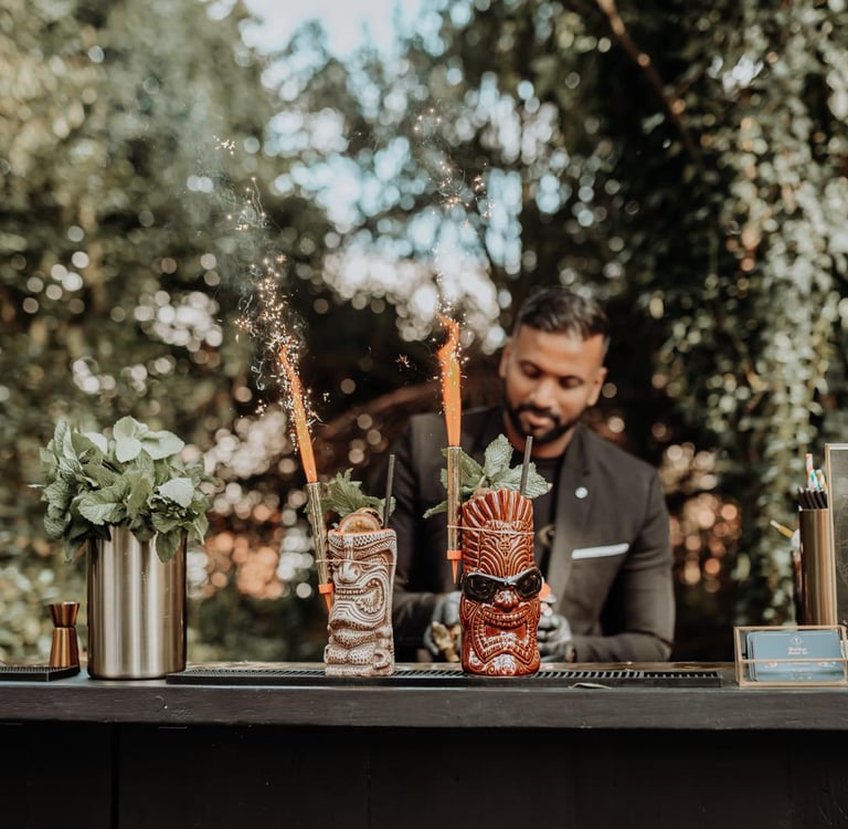 Professional bartender serving tropical tiki cocktails with sparklers at an outdoor garden bar.
