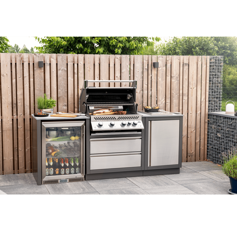 Modern outdoor kitchen with a built-in stainless steel gas grill, mini fridge, and cabinets on a stone patio.