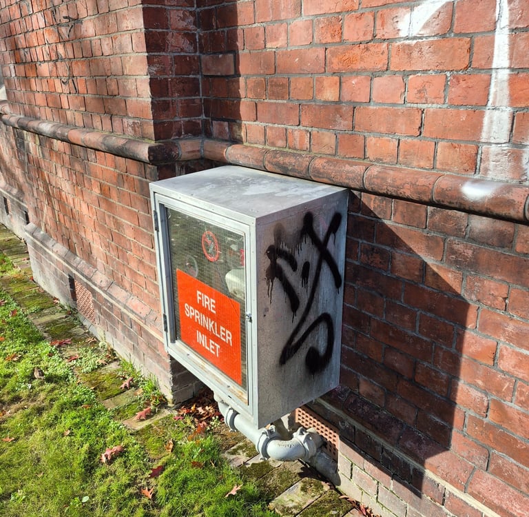 A metal fire sprinkler inlet box with graffiti mounted on an old red brick wall outdoors.