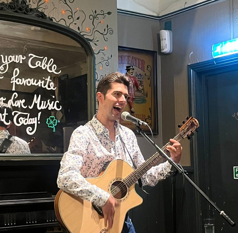 Olly Williams performing live at The Fiddler’s Elbow in Brighton, playing acoustic guitar and singing into a microphone.