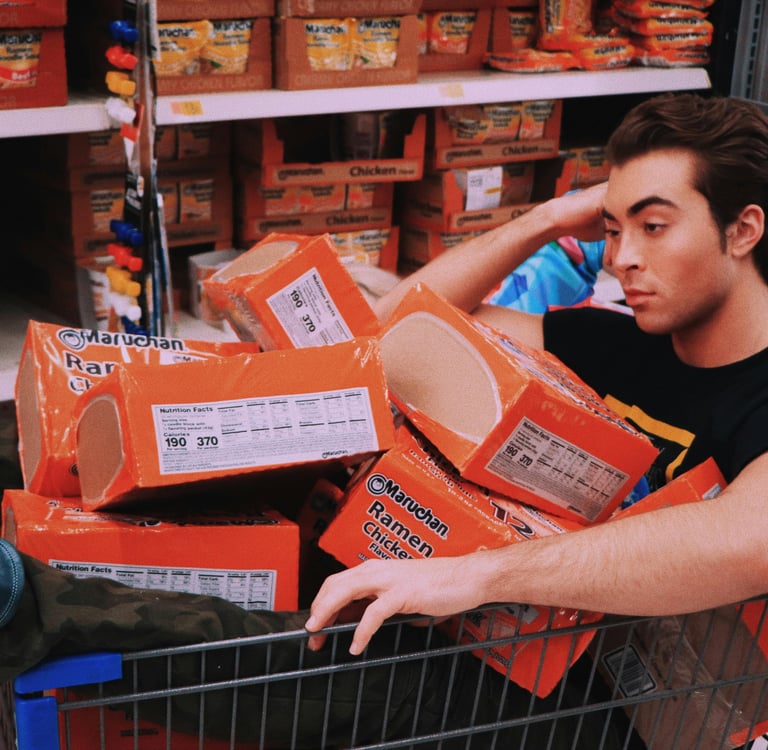 A man sits in a shopping cart filled with orange Maruchan chicken ramen noodle soup boxes in a grocery store aisle.
