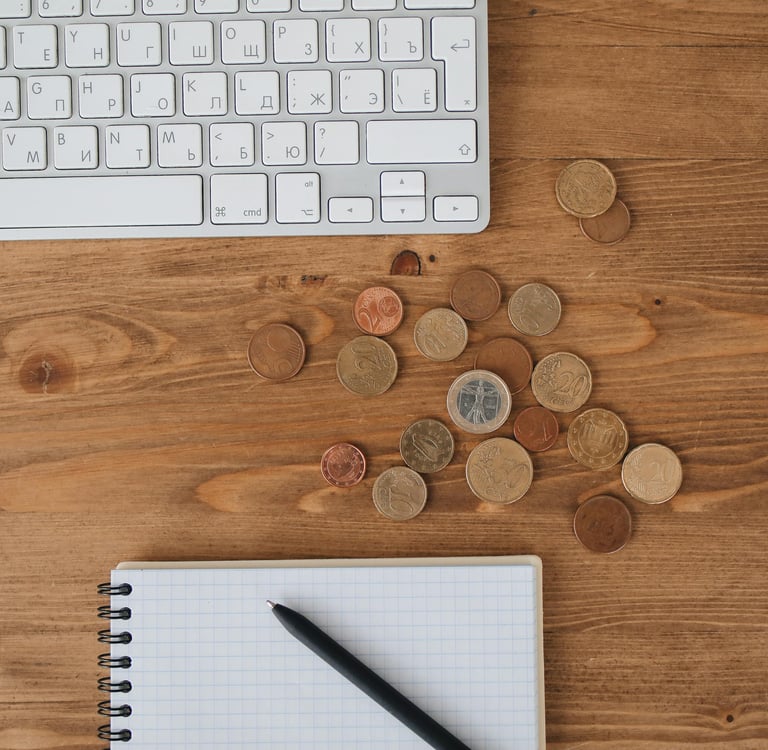 Overhead view of a computer keyboard, Euro coins, and a notebook with a pen on a wooden desk.
