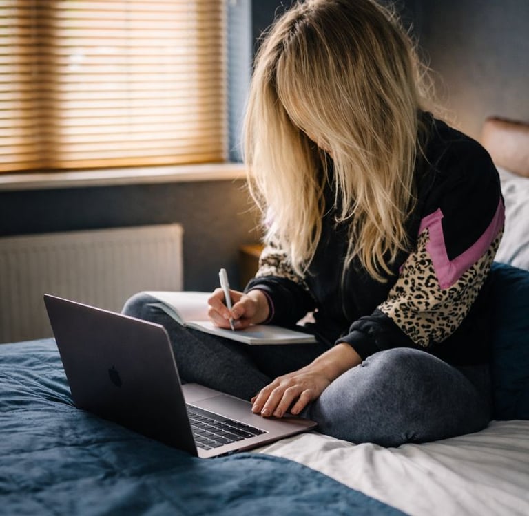 A young woman sits on her bed, using a laptop and notepad
