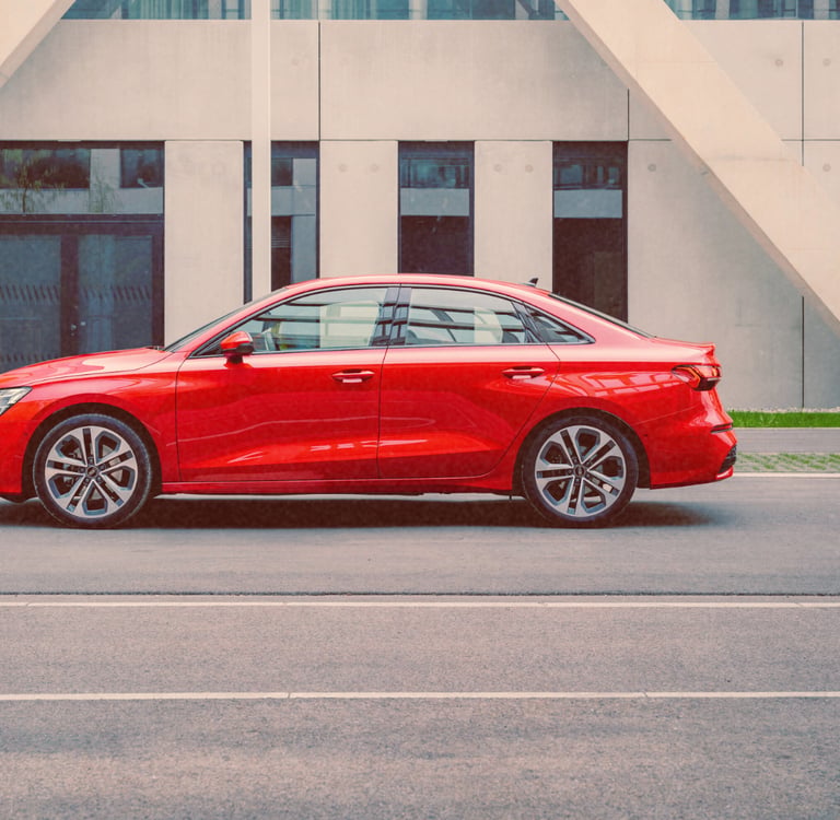 Side profile of an Audi A3 red luxury sedan parked on a city street in front of modern architecture.