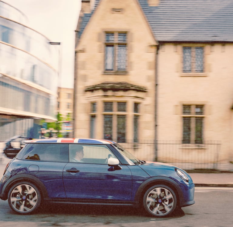 A blue Mini Cooper with a Union Jack roof driving past historic and modern buildings in Oxford.