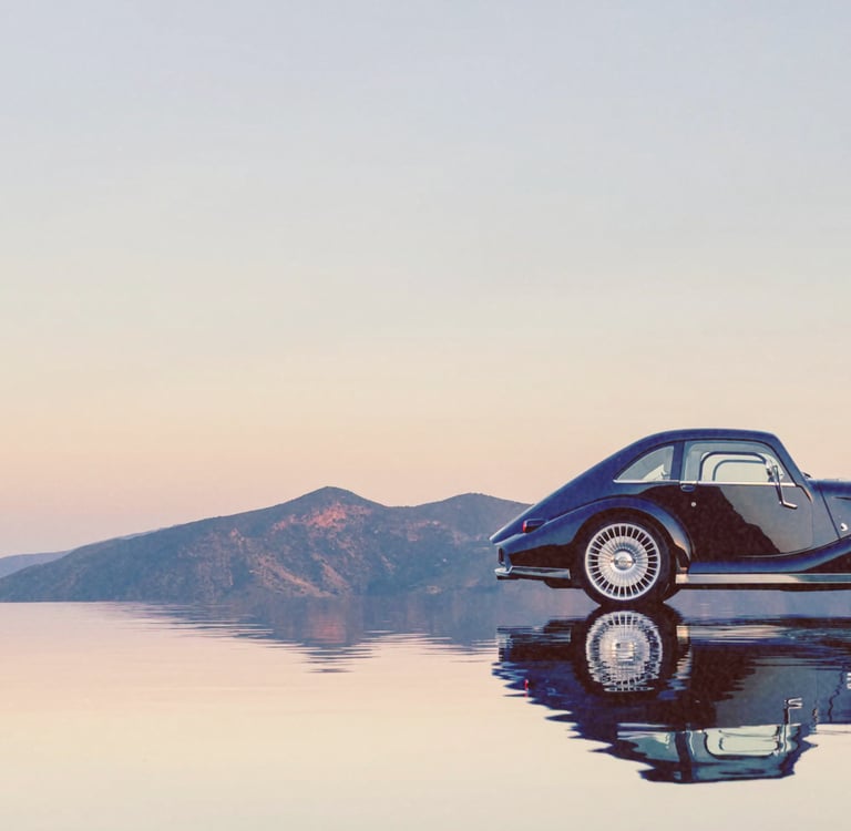 A vintage black luxury car reflecting on a calm water surface with mountains and a sunset sky in the background.