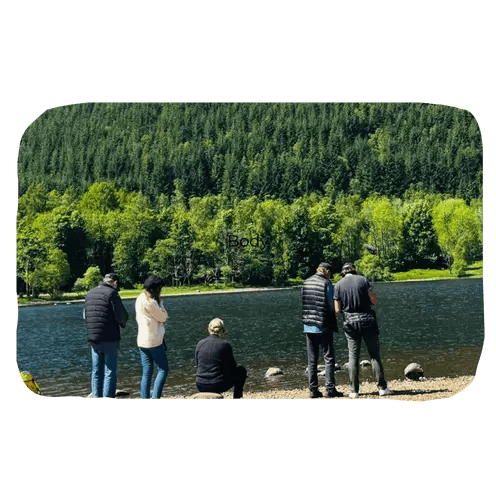 a group of people standing beside a loch with trees across the water