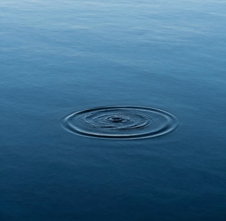 A minimalist, high-angle photograph of a single ripple moving across a perfectly still, deep blue lake. The water is crystalline, reflecting a pale azure sky and absolute stillness.