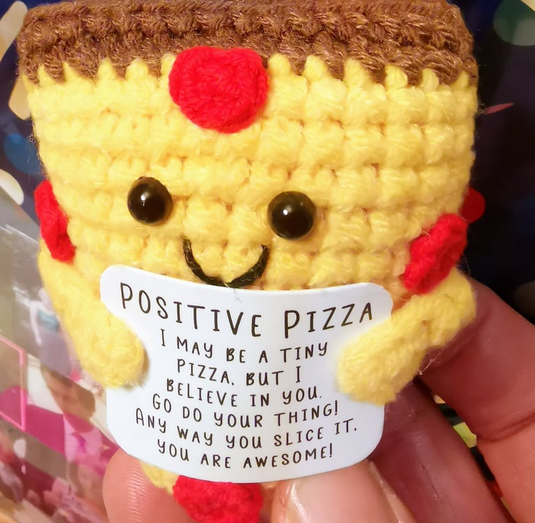 Small yellow crochet pizza amigurumi holding a positive message card with an encouraging quote.