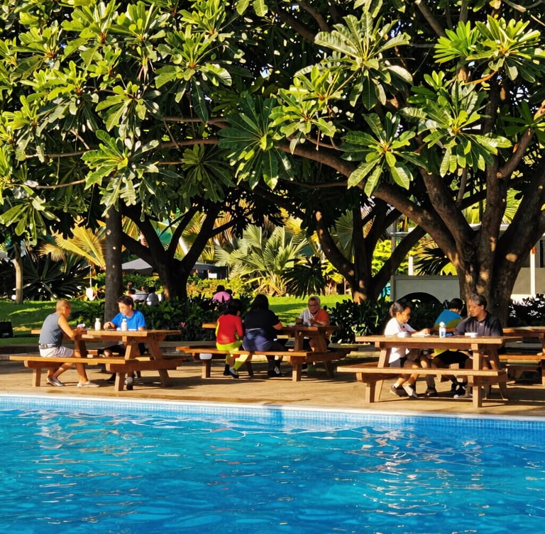 A peaceful shaded picnic area with wooden tables under large leafy trees, families chatting in the background, soft golden sunlight filtering through leaves, vibrant outdoor photography