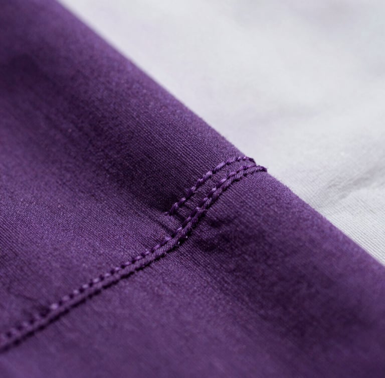 Macro close-up shot of thick 240gsm cotton fabric texture, soft overcast daylight, focus on tight double-needle stitching, deep purple and white tones