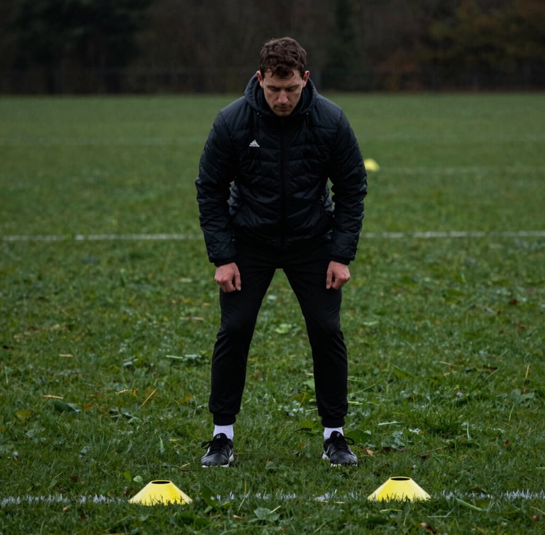 Male football coach setting up agility cones on dew-covered grass under overcast daylight, deep charcoal and grass-green tones, shallow depth of field, moody athletic photography.