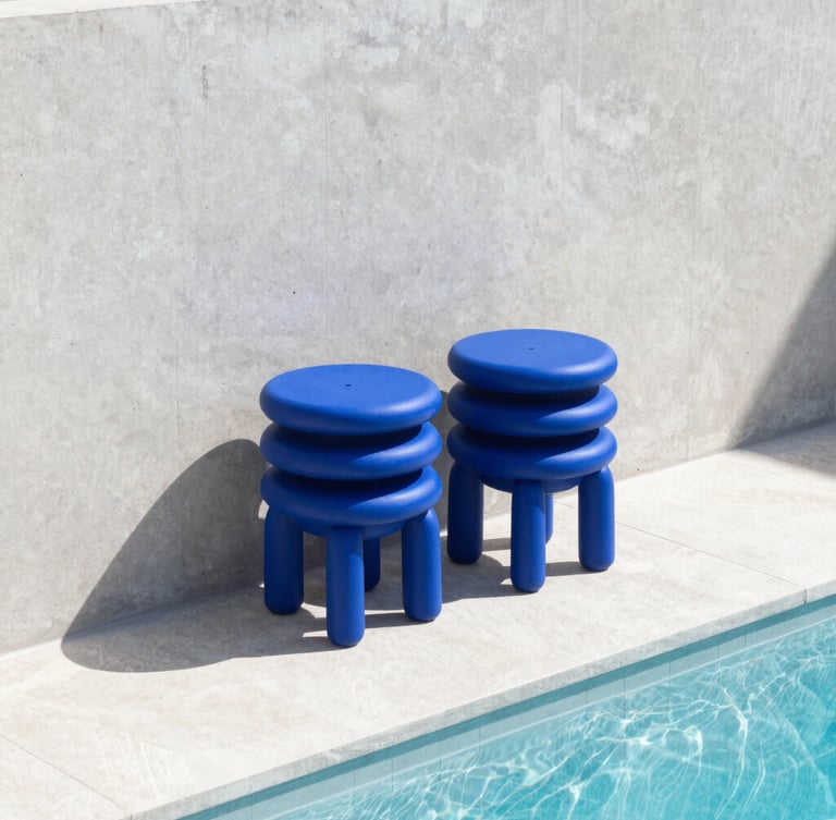 Cobalt blue monolithic resin stools stacked neatly against a sun-drenched concrete wall beside a sparkling pool, hard direct sunlight, sharp graphic shadows, architectural minimalism.