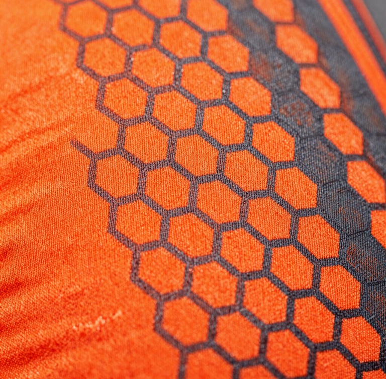 Macro extreme close-up of sublimated athletic fabric weave, showing vibrant neon-orange and charcoal hexagonal patterns integrated into the technical fibers, sharp studio lighting, 85mm lens