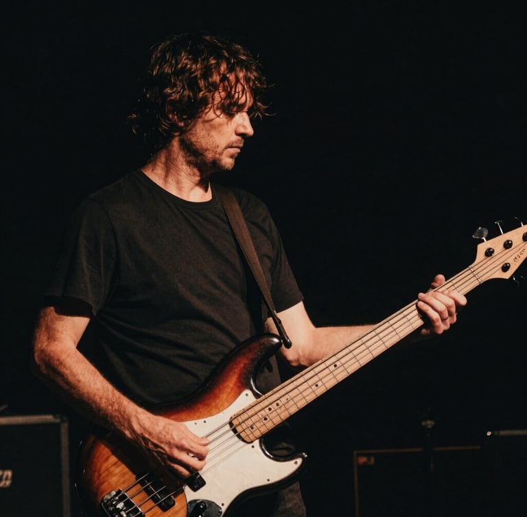 Moody low-key photography of Pierre-Yves Clarence holding his electric bass, high contrast stage lighting, deep shadows, warm amber undertones, 35mm film grain.