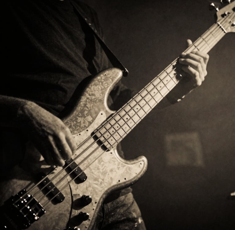 Close-up of bassist hands on the strings of an electric bass, dramatic side lighting, high contrast monochrome, warm amber undertones, smoky jazz club atmosphere.