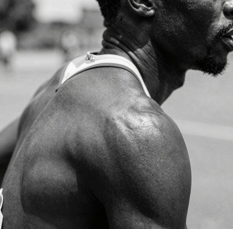 High-contrast black and white close-up of a Ghanaian athlete's shoulder in motion, hard direct sunlight casting deep shadows, raw skin texture.