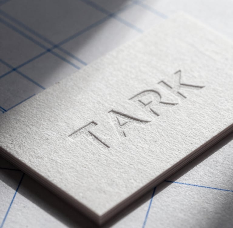 Stark macro photography of a physical embossed business card on heavy cotton paper, sharp directional studio light casting deep shadows, technical blue grid lines faintly visible.