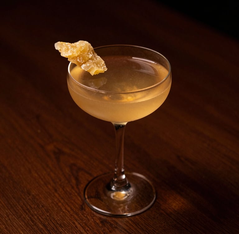 A minimalist cocktail in a delicate coupe glass, garnished with a single slice of crystallized ginger, warm amber candlelight glow, deep shadows