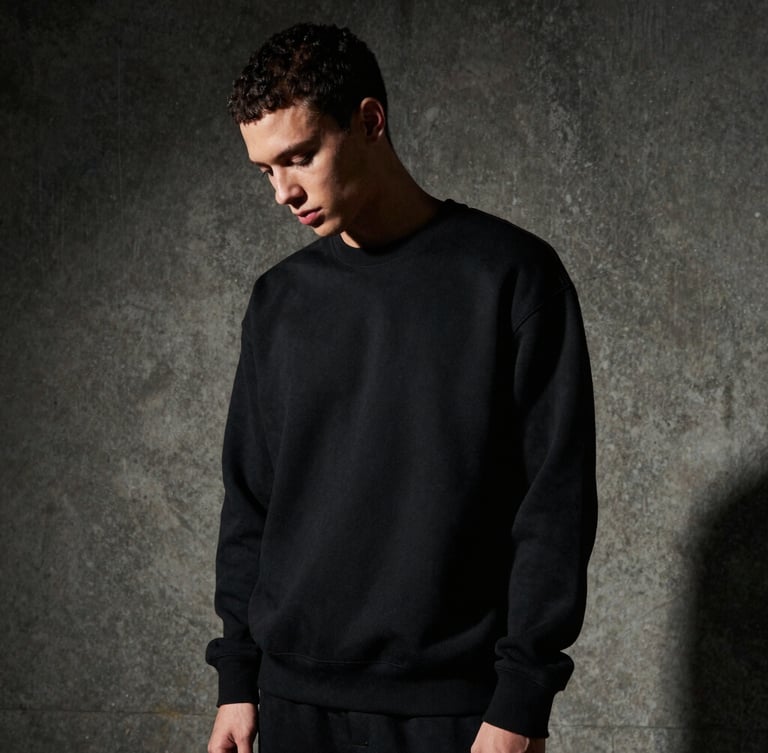 Stoic model in a heavyweight black structured sweatshirt looking down, dramatic side-lighting casting deep shadows, raw concrete background, 35mm film grain