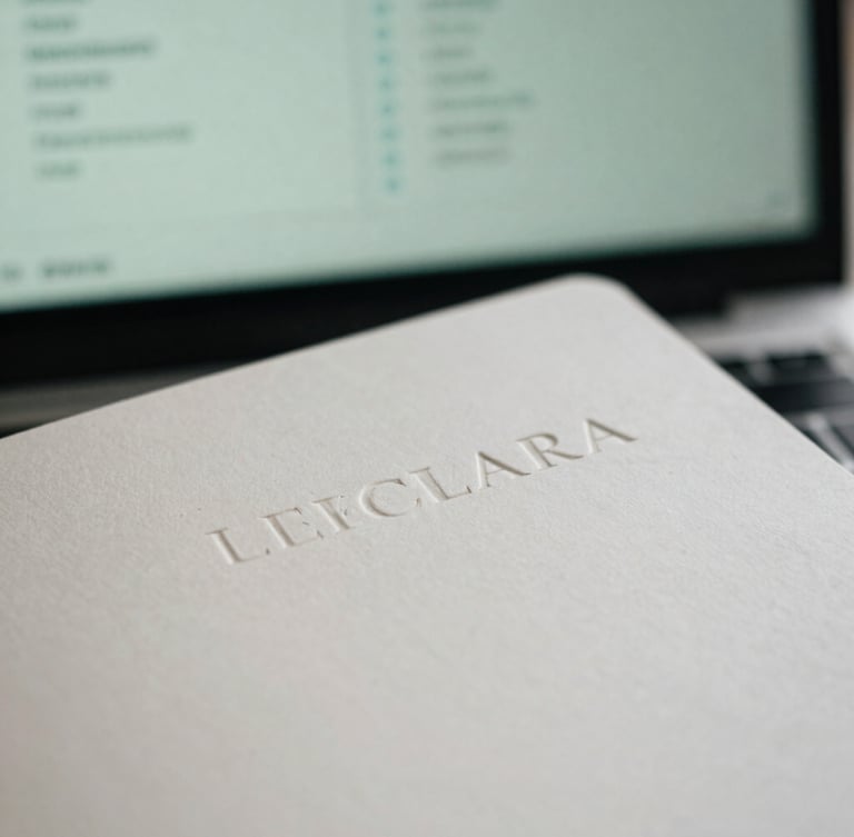Close-up of an embossed LECLARA brand asset (e.g., stationery) on a clean desktop, with a blurred analytical dashboard in the background, natural overcast London daylight, soft shadows, macro detail.
