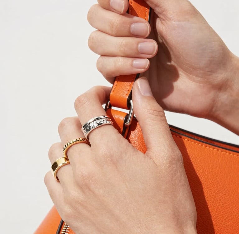 Macro close-up of dynamic hands wearing multiple textured silver and gold rings, holding a vibrant orange leather bag, bright high-key fashion photography, crisp natural daylight, sharp macro details, 35mm lens