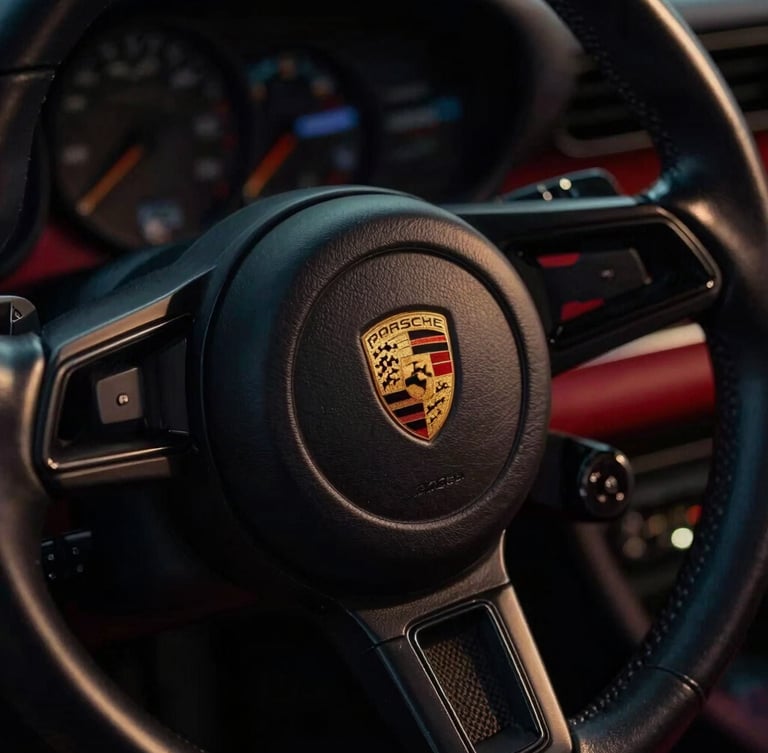Extreme close-up of a classic Porsche 911 leather steering wheel, gold crest emblem sharply in focus, low-key dramatic lighting, deep shadows, premium editorial style.