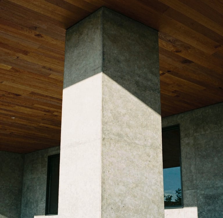 Tight geometric crop of a raw board-formed concrete pillar meeting a high timber ceiling, sharp natural daylight casting deep high-contrast shadows, architectural film photography style.