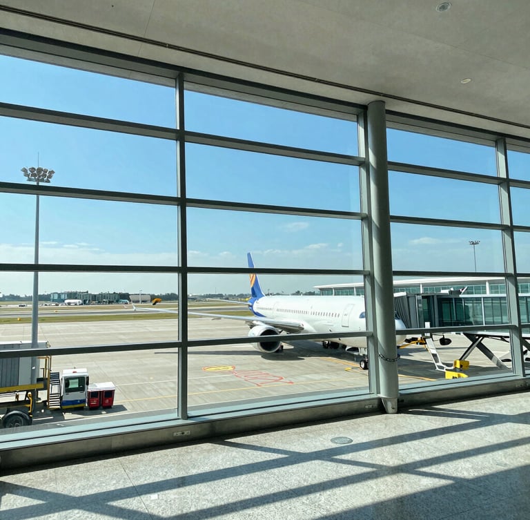 Wide environmental shot of a modern clean airport terminal with bright natural daylight streaming through giant glass windows, a commercial airplane visible on the tarmac